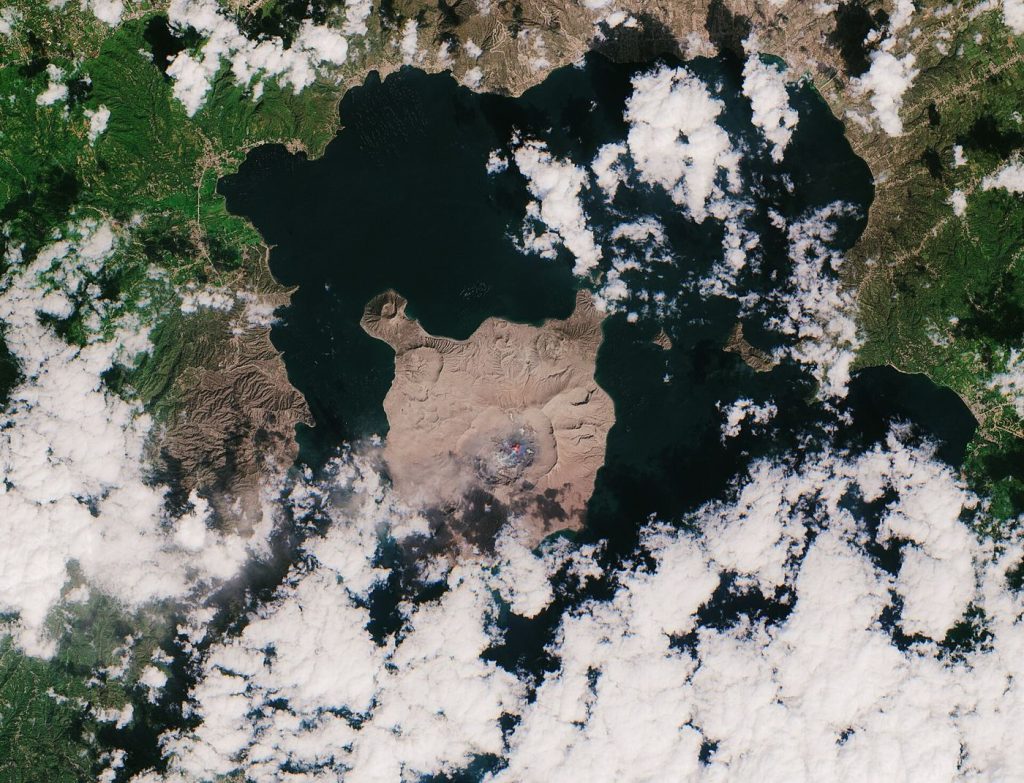 Island Volcano Ash Plume Reshapes Local Air Routes A volcanic eruption on an island sending a thick grey ash cloud into the sky above the coast.